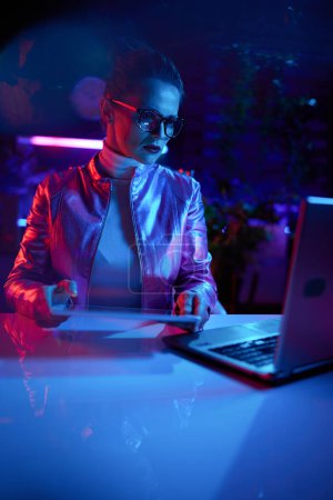Téléchargez les photos : Neon metaverse concept futuriste. élégante femelle dans des lunettes avec ordinateur portable en utilisant tablette PC dans le bureau moderne. - en image libre de droit