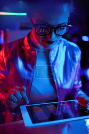 Photo for Neon metaverse futuristic concept. trendy female in glasses with tablet PC and charts reflecting in glasses in modern office. - Royalty Free Image