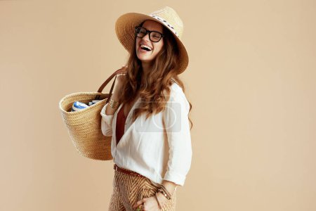 Téléchargez les photos : Vacances à la plage. femme au foyer moderne souriante en chemisier blanc et short isolé sur beige en lunettes avec sac en paille et chapeau en paille. - en image libre de droit
