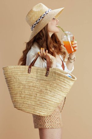 Photo for Beach vacation. happy modern middle aged woman in white blouse and shorts against beige background with straw bag, carrot juice and straw hat. - Royalty Free Image