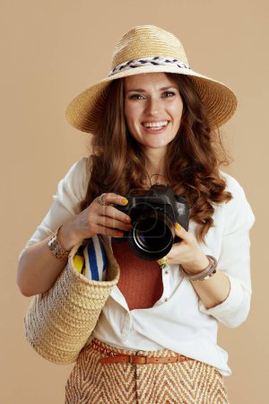 Téléchargez les photos : Vacances à la plage. femme souriante et tendance de 40 ans en chemisier blanc et short sur fond beige avec sac en paille, appareil photo et chapeau d'été. - en image libre de droit