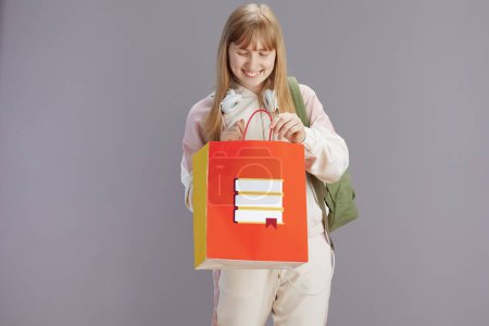 Foto de Feliz chica adolescente moderna en chándal beige con mochila, bolsa de compras y auriculares aislados en gris. - Imagen libre de derechos