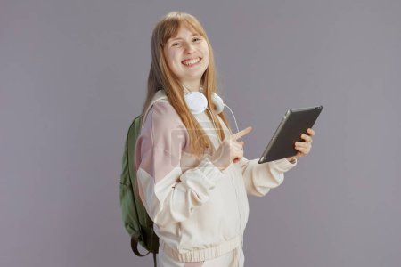 Foto de Pupila moderna sonriente en chándal beige con mochila, auriculares y tableta digital aislada sobre fondo gris. - Imagen libre de derechos