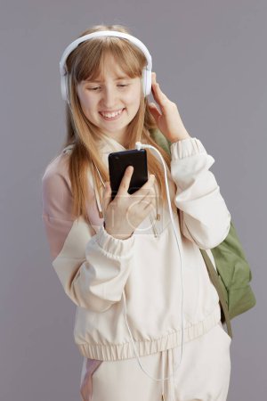 Photo for Happy trendy girl in beige tracksuit with backpack listening to the music with headphones and using smartphone applications isolated on grey background. - Royalty Free Image