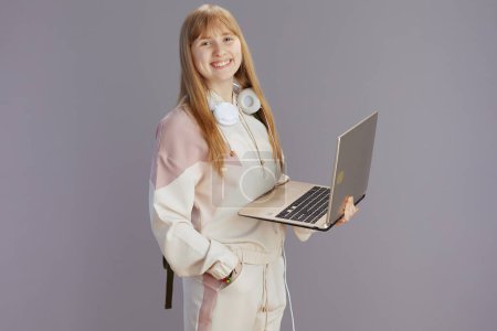 Photo for Portrait of happy trendy teen girl in beige tracksuit with headphones using laptop isolated on grey background. - Royalty Free Image