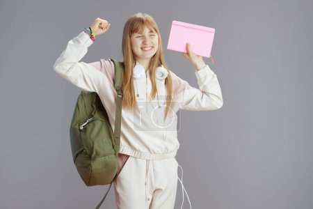 Foto de Sonriente chica adolescente moderna en chándal beige con mochila, libros de trabajo y auriculares contra el regocijo gris. - Imagen libre de derechos