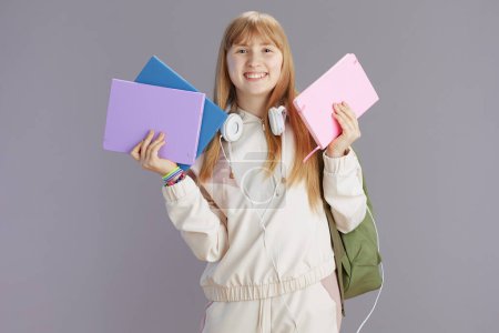 Photo for Portrait of smiling modern pupil in beige tracksuit with backpack, workbooks and headphones isolated on grey background. - Royalty Free Image
