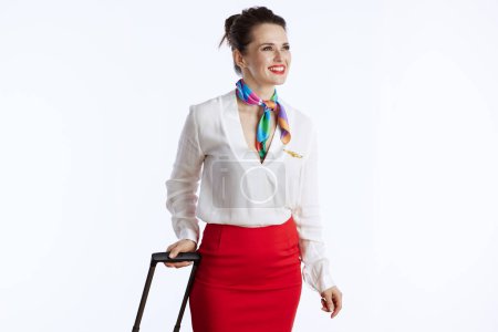 Foto de Asistente de vuelo femenino elegante sonriente contra fondo blanco en uniforme con bolsa de viaje mirando a la distancia. - Imagen libre de derechos