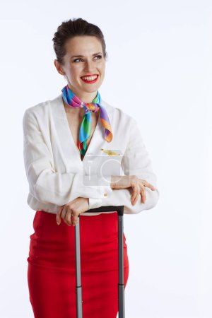 Foto de Pensativo elegante azafata de aire femenino contra fondo blanco en uniforme con bolsa de carro mirando a la distancia. - Imagen libre de derechos