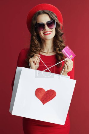 Téléchargez les photos : Joyeux Valentin. heureuse élégante femme de 40 ans en robe rouge et béret sur fond rouge avec carte de crédit et sac à provisions et coeur. - en image libre de droit