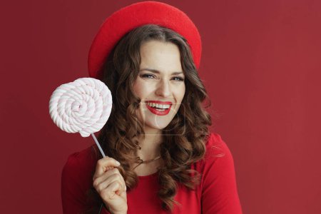 Foto de Feliz San Valentín. mujer elegante feliz en vestido rojo y boina con caramelo en palo aislado en rojo. - Imagen libre de derechos