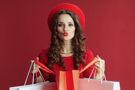 Foto de Feliz San Valentín. mujer elegante con el pelo ondulado largo en vestido rojo y boina sobre fondo rojo con bolsas de compras enviando un beso. - Imagen libre de derechos