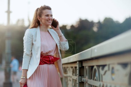 Téléchargez les photos : Souriant élégante femme de 40 ans en robe rose et veste blanche dans la ville parlant sur un smartphone sur le pont. - en image libre de droit