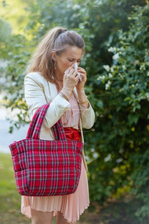 Téléchargez les photos : Femelle tendance en robe rose et veste blanche dans la ville avec serviette et sac rouge a une attaque d'allergie. - en image libre de droit