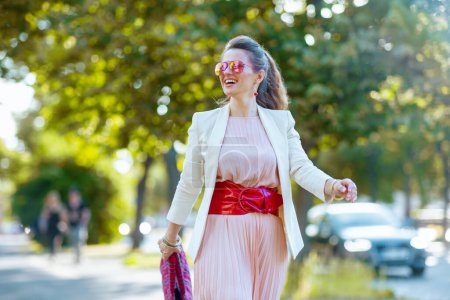 Téléchargez les photos : Femme moderne souriante en robe rose et veste blanche dans la ville avec des lunettes de soleil marche. - en image libre de droit