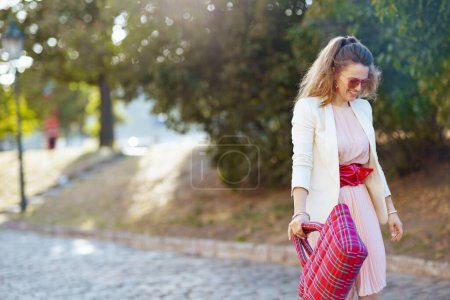 Téléchargez les photos : Heureuse femme élégante en robe rose et veste blanche dans la ville avec sac rouge et lunettes de soleil marche. - en image libre de droit