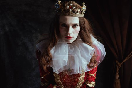 Téléchargez les photos : Effrayante reine médiévale en robe rouge avec col blanc et couronne sur fond gris foncé. - en image libre de droit