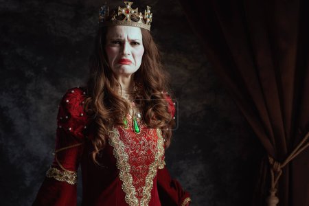 Photo for Unhappy medieval queen in red dress with white makeup and crown on dark gray background. - Royalty Free Image