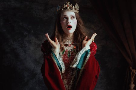 Foto de Impactada reina medieval en vestido rojo con maquillaje blanco y corona sobre fondo gris oscuro. - Imagen libre de derechos