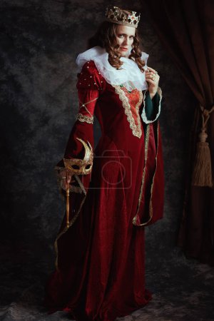 Photo for Full length portrait of insidious medieval queen in red dress with venetian mask, white collar and crown on dark gray background. - Royalty Free Image