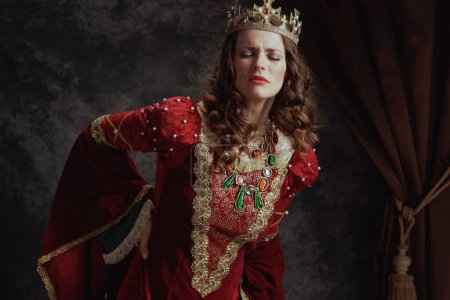Foto de Cansada reina medieval en vestido rojo con corona con dolor de espalda sobre fondo gris oscuro. - Imagen libre de derechos