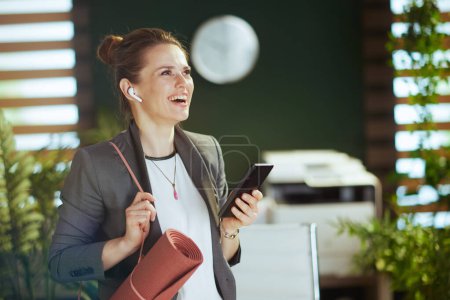 Foto de Lugar de trabajo sostenible. mujer de negocios moderna sonriente en la oficina verde moderna con auriculares inalámbricos y esterilla de yoga utilizando la aplicación para teléfonos inteligentes. - Imagen libre de derechos