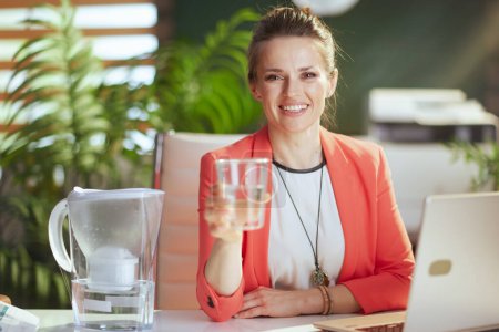 Téléchargez les photos : Un lieu de travail durable. Portrait de femme d'affaires heureuse et moderne au travail dans une veste rouge avec tasse d'eau, filtre à eau et ordinateur portable. - en image libre de droit
