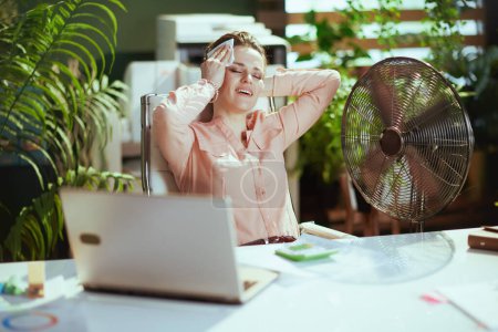 Photo for Sustainable workplace. happy modern 40 years old business woman at work with electric fan, laptop and napkin. - Royalty Free Image