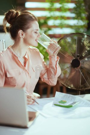 Photo for Sustainable workplace. smiling modern female employee at work with electric fan, laptop, cup of water and lemon. - Royalty Free Image