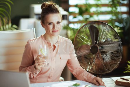 Foto de Lugar de trabajo sostenible. sonriente trabajadora moderna en moderna oficina verde con ventilador eléctrico, taza de agua y limón. - Imagen libre de derechos