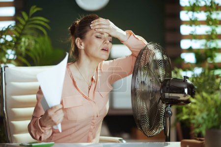 Photo for Sustainable workplace. modern 40 years old woman employee at work with documents and electric fan suffering from summer heat. - Royalty Free Image