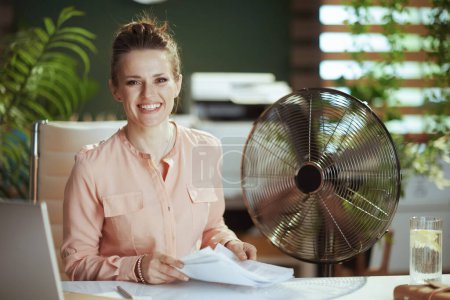 Photo for Sustainable workplace. Portrait of smiling modern 40 years old small business owner woman in modern green office with documents and electric fan. - Royalty Free Image