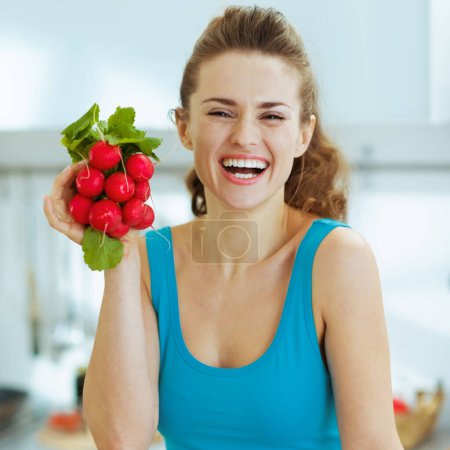 Foto de Mujer joven sonriente sosteniendo racimo de rábanos en la cocina - Imagen libre de derechos