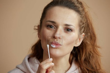 Foto de Mujer de 40 años con herpes en los labios aplicando ungüento con hisopo de algodón sobre fondo beige. - Imagen libre de derechos