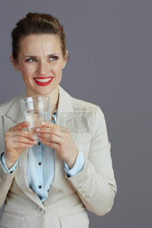 Téléchargez les photos : Souriant femme d'affaires d'âge moyen à la mode dans un costume d'affaires léger avec verre d'eau sur fond gris. - en image libre de droit