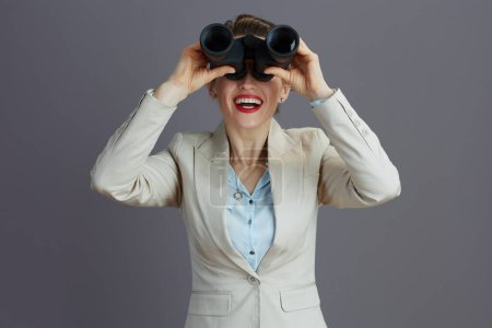 Photo for Smiling trendy 40 years old woman employee in a light business suit with binoculars isolated on grey. - Royalty Free Image