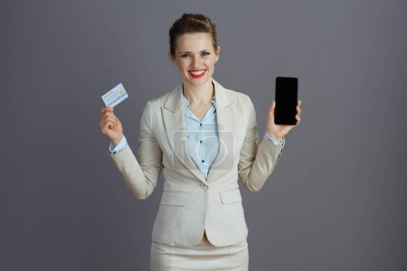 Foto de Empleada femenina moderna sonriente en un traje de negocios ligero con teléfono inteligente y tarjeta de crédito contra fondo gris. - Imagen libre de derechos