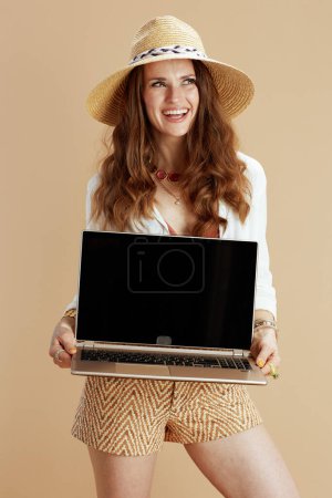 Photo for Beach vacation. smiling elegant middle aged woman in white blouse and shorts isolated on beige background with straw hat showing laptop blank screen. - Royalty Free Image