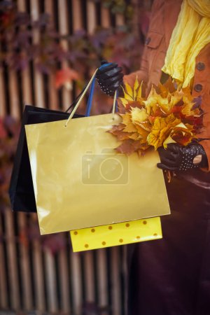 Foto de Hola octubre. Primer plano de la mujer de mediana edad en gabardina naranja con bolsas de compras y hojas amarillas de otoño en la ciudad. - Imagen libre de derechos