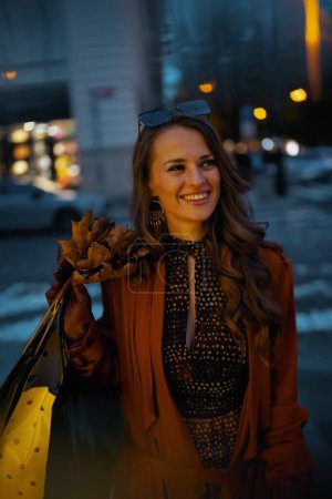 Foto de Hola octubre. Sonriente hembra de moda en gabardina naranja con bolsas de compras y hojas amarillas otoñales en la ciudad. - Imagen libre de derechos