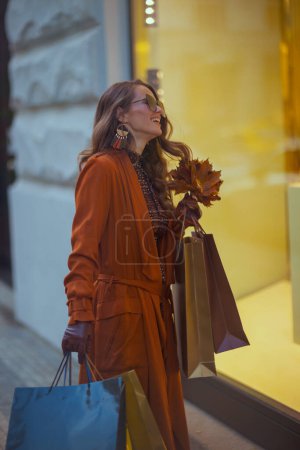 Foto de Hola septiembre. mujer de mediana edad feliz en gabardina naranja con bolsas de compras y hojas amarillas de otoño cerca de la tienda en la ciudad. - Imagen libre de derechos