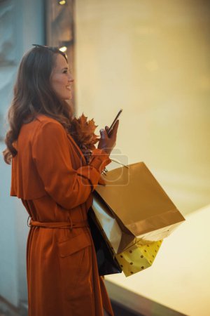 Téléchargez les photos : Bonjour, novembre. heureuse femme élégante en trench-coat orange avec sacs à provisions, smartphone et feuilles jaune automne près du magasin dans la ville. - en image libre de droit