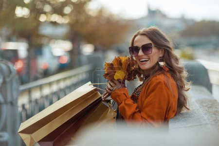 Hello september. smiling modern woman in brown trench coat with shopping bags and autumn yellow leaves in the city.