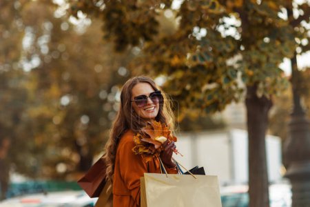 Téléchargez les photos : Bonjour octobre. heureuse femme à la mode en trench-coat marron avec des sacs à provisions et des feuilles jaune automne dans la ville. - en image libre de droit