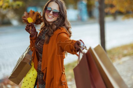 Foto de Hola otoño. mujer con estilo feliz en gabardina marrón con bolsas de compras y hojas amarillas de otoño en la ciudad. - Imagen libre de derechos
