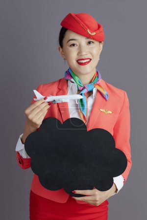 Photo for Happy elegant asian female air hostess in red skirt, jacket and hat uniform with a little airplane showing blank cloud shape board against gray background. - Royalty Free Image