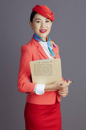Photo for Smiling modern asian female stewardess in red skirt, jacket and hat uniform with clipboard isolated on grey. - Royalty Free Image