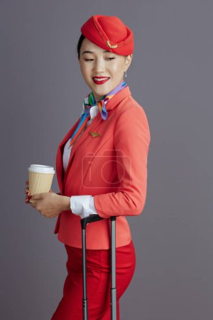 Foto de Pensativo elegante aire azafata asiático mujer en rojo falda, chaqueta y sombrero uniforme con café taza y carro bolsa aislado en gris fondo. - Imagen libre de derechos