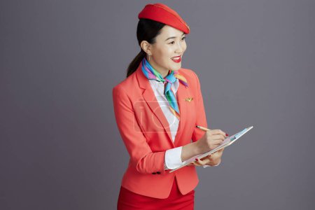 Foto de Sonriente elegante aire azafata asiático mujer en rojo falda, chaqueta y sombrero uniforme con portapapeles sobre gris fondo. - Imagen libre de derechos