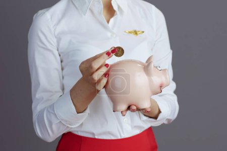 Photo for Closeup on modern asian female air hostess in red skirt and hat uniform with piggy bank isolated on grey background. - Royalty Free Image